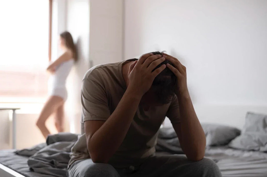 Ein Mann sitzt verzweifelt auf dem Bett, hält seinen Kopf in den Händen. Im Hintergrund steht eine Frau mit verschränkten Armen, unscharf im Bild.
