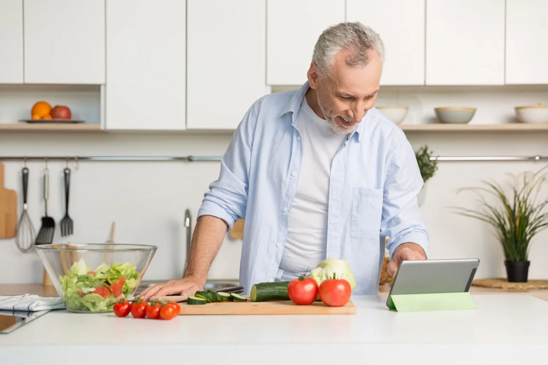 Älterer Mann bereitet in einer modernen Küche einen frischen Salat zu und schaut dabei auf ein Tablet.