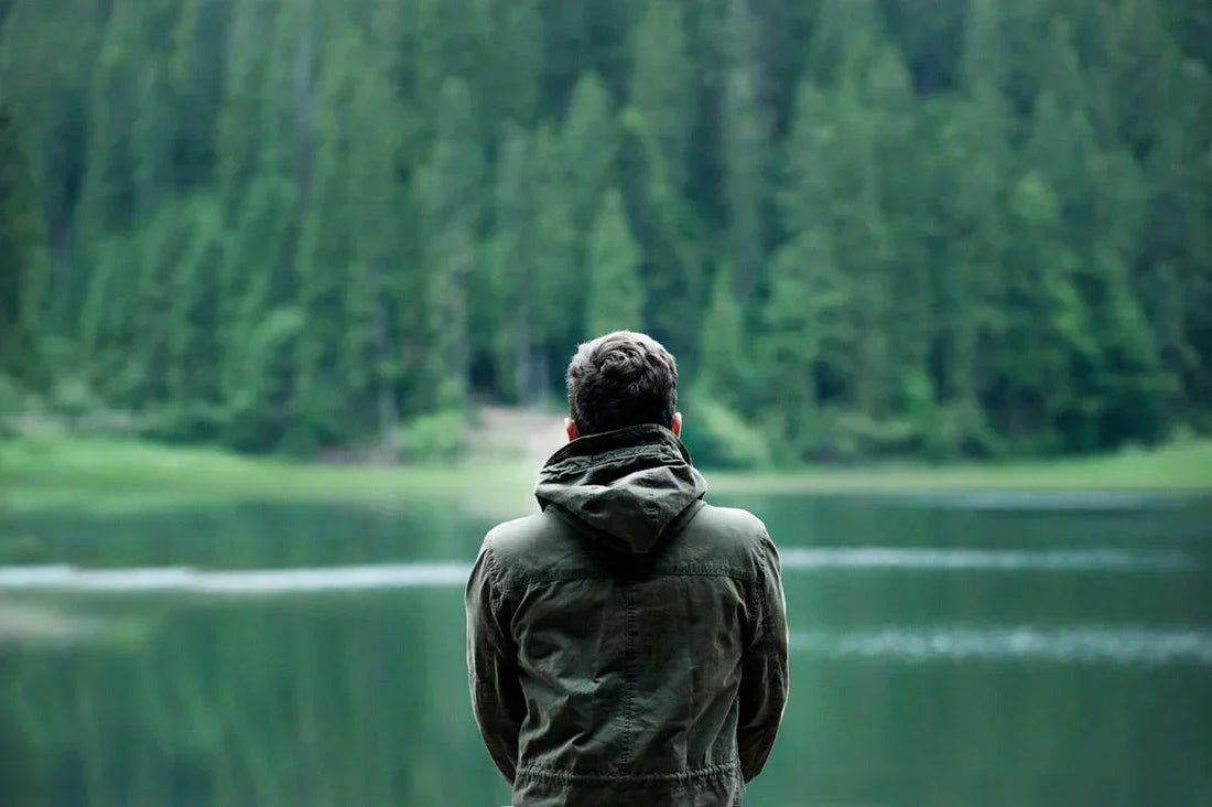 Mann in grüner Jacke steht mit dem Rücken zur Kamera und blickt auf einen ruhigen See, umgeben von dichten, grünen Wäldern.