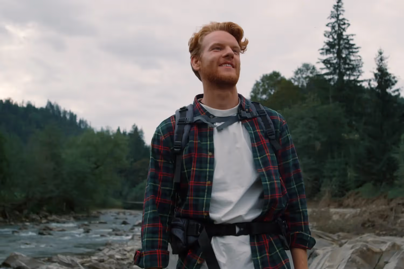 Ein Mann steht in der Natur und genießt die Umgebung. Man sieht einen Fluss und Bäume im Hintergrund.