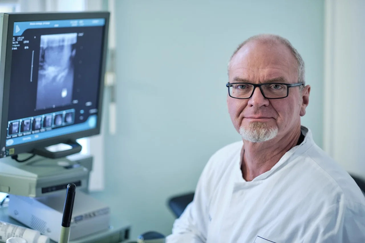 Prof. Dr. Michael Zitzmann sitzt in seiner Praxis, trägt einen weißen Kittel und blickt professionell in die Kamera. Neben ihm ein Ultraschallgerät mit Monitor.
