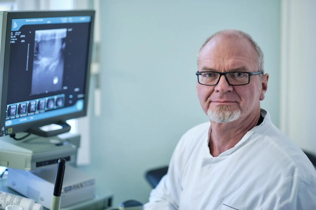 Prof. Dr. Michael Zitzmann sitzt in seiner Praxis, trägt einen weißen Kittel und blickt professionell in die Kamera. Neben ihm ein Ultraschallgerät mit Monitor.