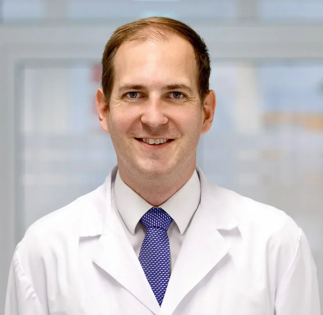 Oberarzt Dr. Wessels in weißem Arztkittel und Krawatte, freundlich lächelnd vor einem hellen, modernen Hintergrund.