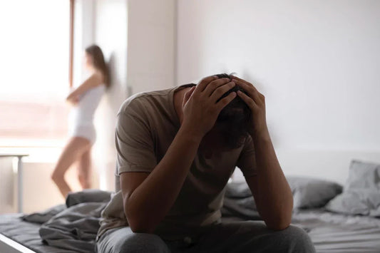 Ein Mann sitzt verzweifelt auf dem Bett, hält seinen Kopf in den Händen. Im Hintergrund steht eine Frau mit verschränkten Armen, unscharf im Bild.