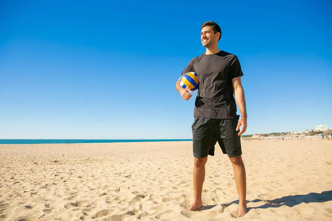 Mann steht barfuß am sonnigen Sandstrand mit Volleyball in der Hand – Symbolbild für Bewegung an der frischen Luft und natürliche Vitamin-D-Produktion durch Sonnenlicht.