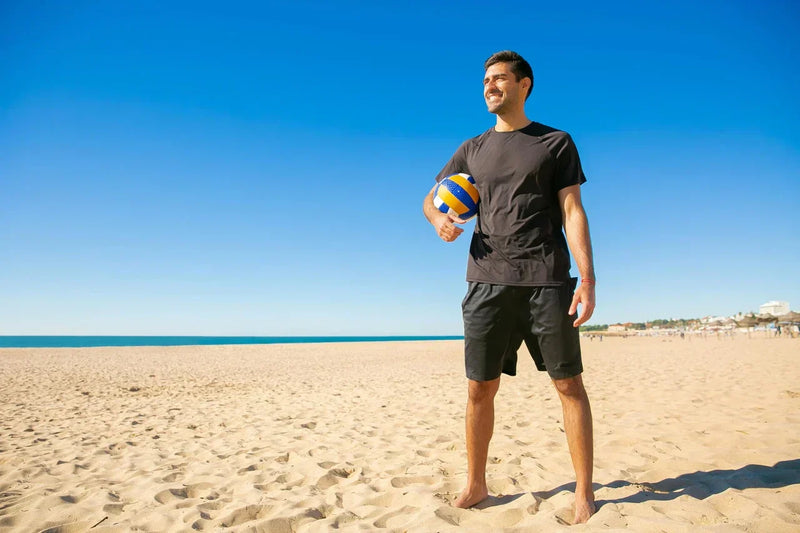 Mann steht barfuß am sonnigen Sandstrand mit Volleyball in der Hand – Symbolbild für Bewegung an der frischen Luft und natürliche Vitamin-D-Produktion durch Sonnenlicht.