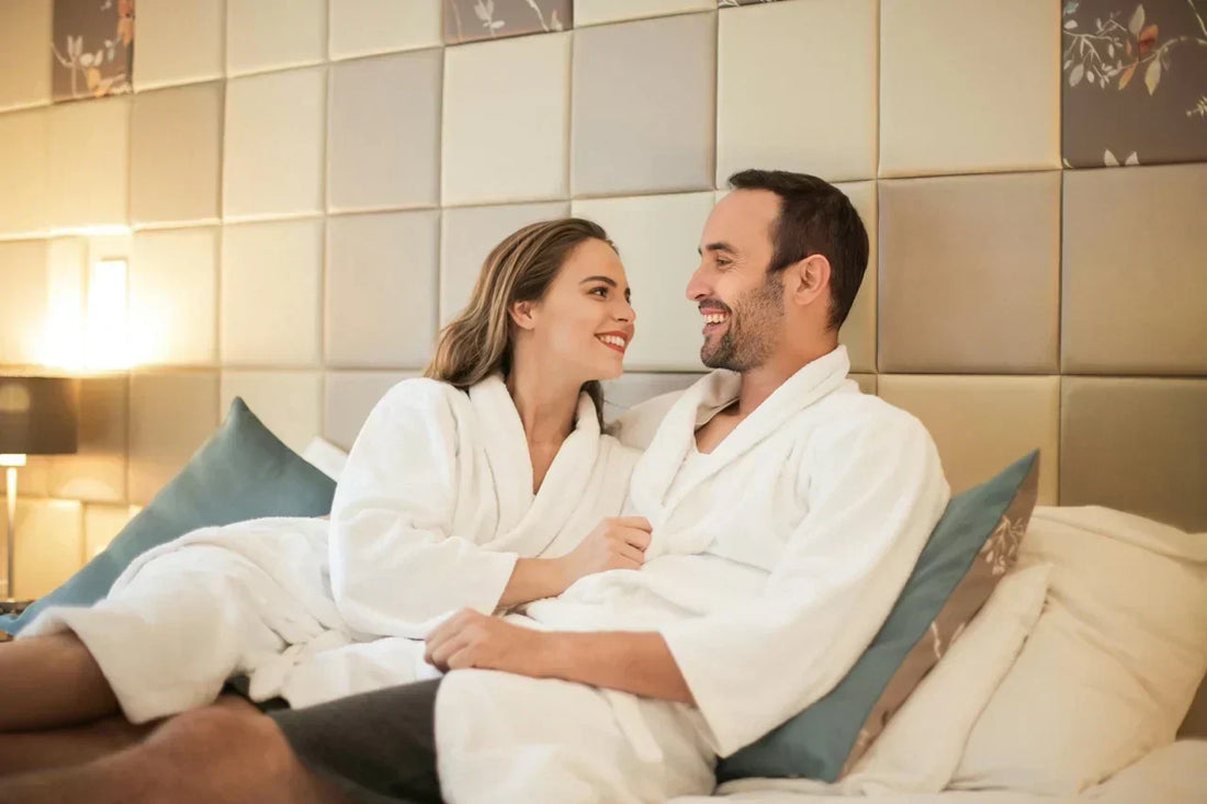 Ein lächelndes Paar in Bademänteln liegt entspannt auf dem Bett und schaut sich liebevoll an.