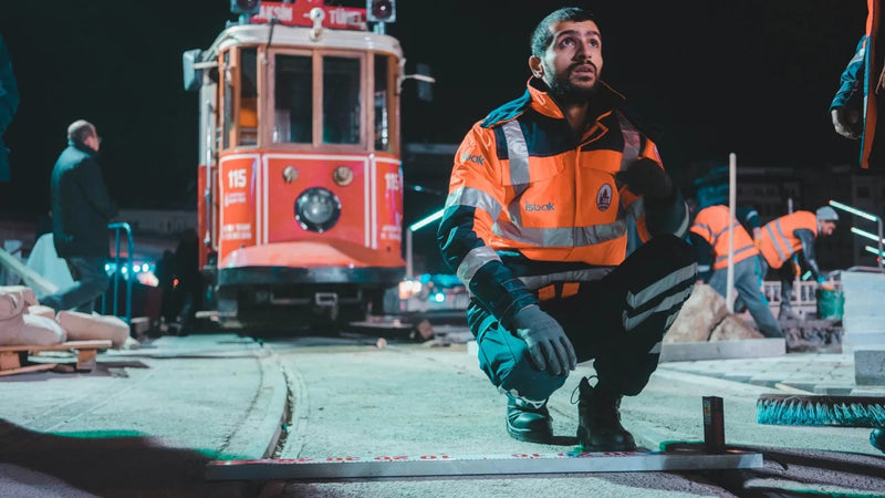 Mann in Schutzkleidung arbeitet nachts auf einer Baustelle im Gleisbereich, kniet auf dem Boden und misst eine Schiene, im Hintergrund steht eine Straßenbahn und weitere Arbeiter führen Bauarbeiten aus.