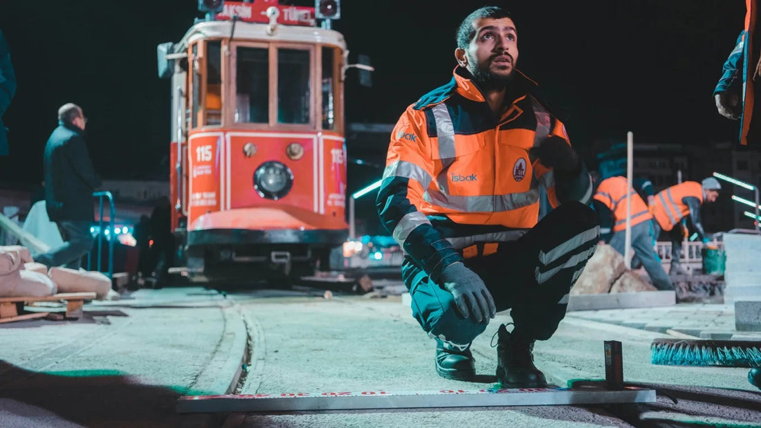 Mann in Schutzkleidung arbeitet nachts auf einer Baustelle im Gleisbereich, kniet auf dem Boden und misst eine Schiene, im Hintergrund steht eine Straßenbahn und weitere Arbeiter führen Bauarbeiten aus.