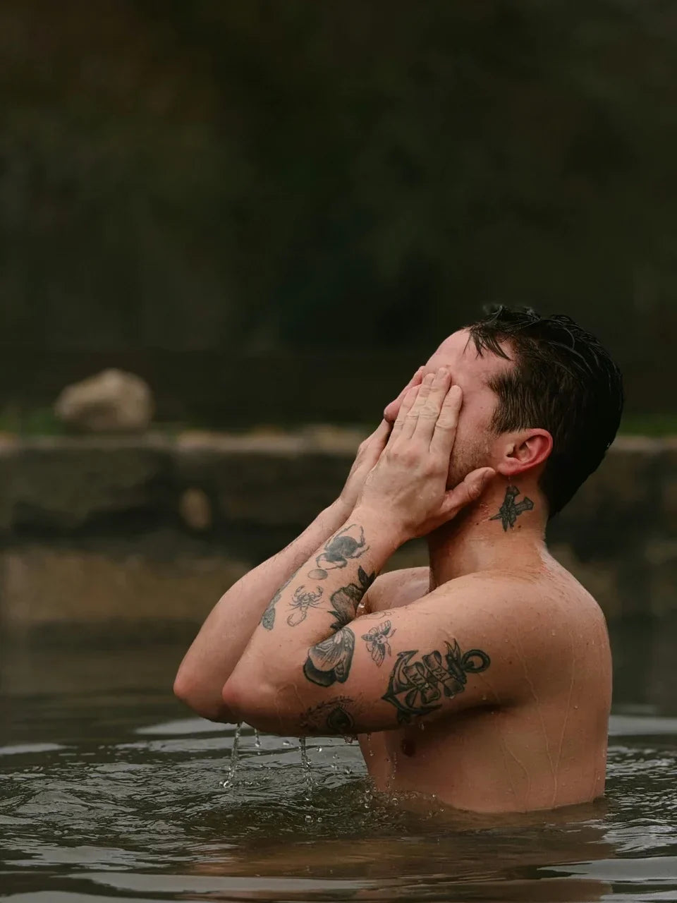 Mann mit Tattoos steht bis zur Brust im Wasser eines Sees und wäscht sich das Gesicht, umgeben von Natur.