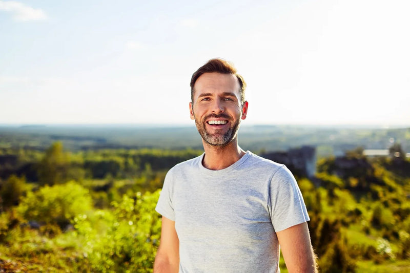 Ein lächelnder Mann mit kurzem Bart und braunen Haaren steht in der Natur. Er trägt ein hellgraues T-Shirt, im Hintergrund eine weite Landschaft mit grünen Bäumen und Hügeln.