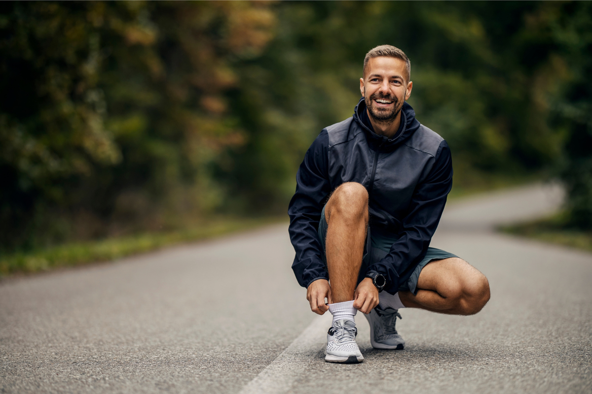 Lächelnder Mann in Laufkleidung hockt auf einer Landstraße im Wald und macht sich bereit zum Joggen